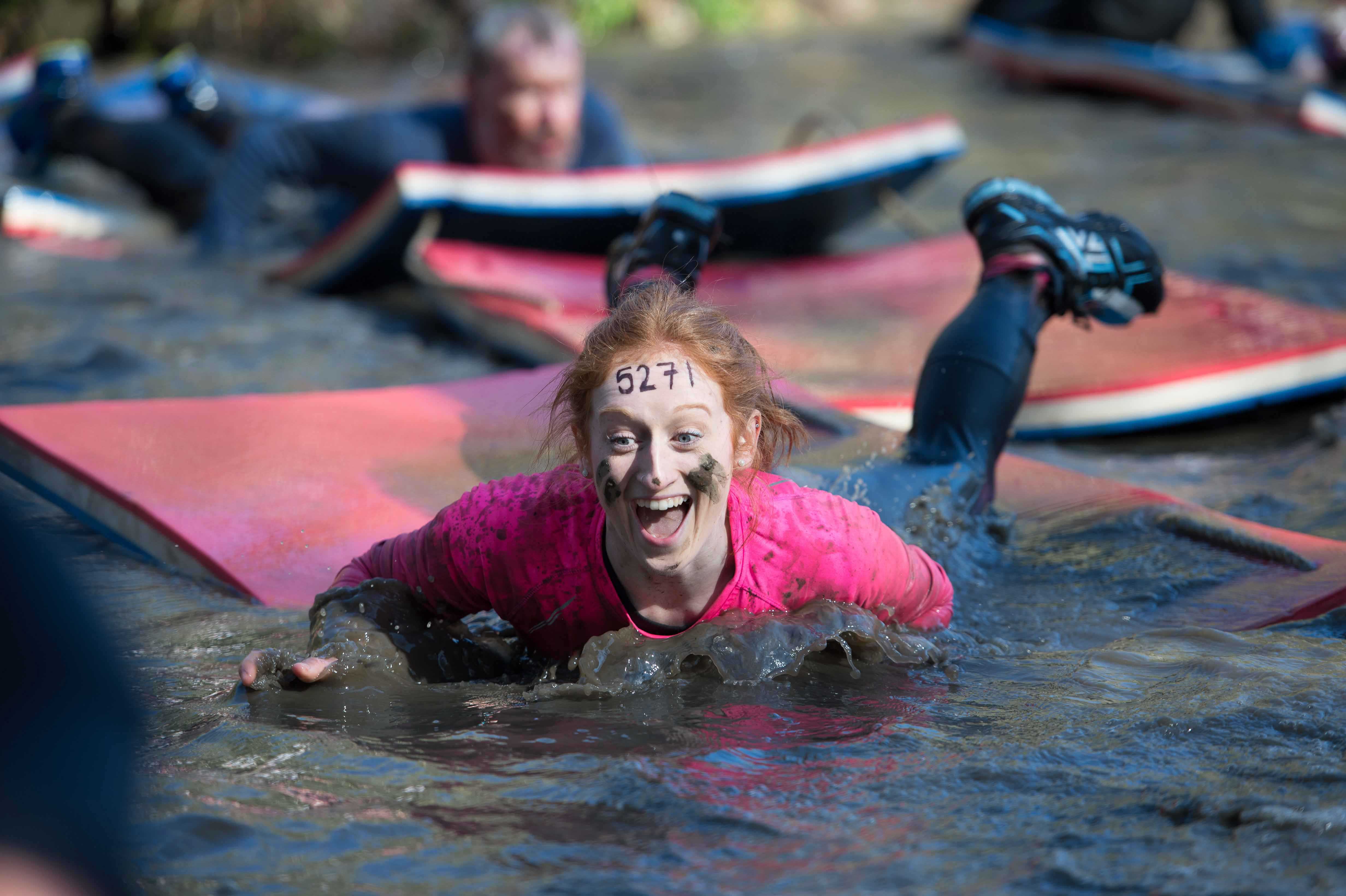 A cheerful young woman smiling while she navigates a muddy bank using a floating mattress