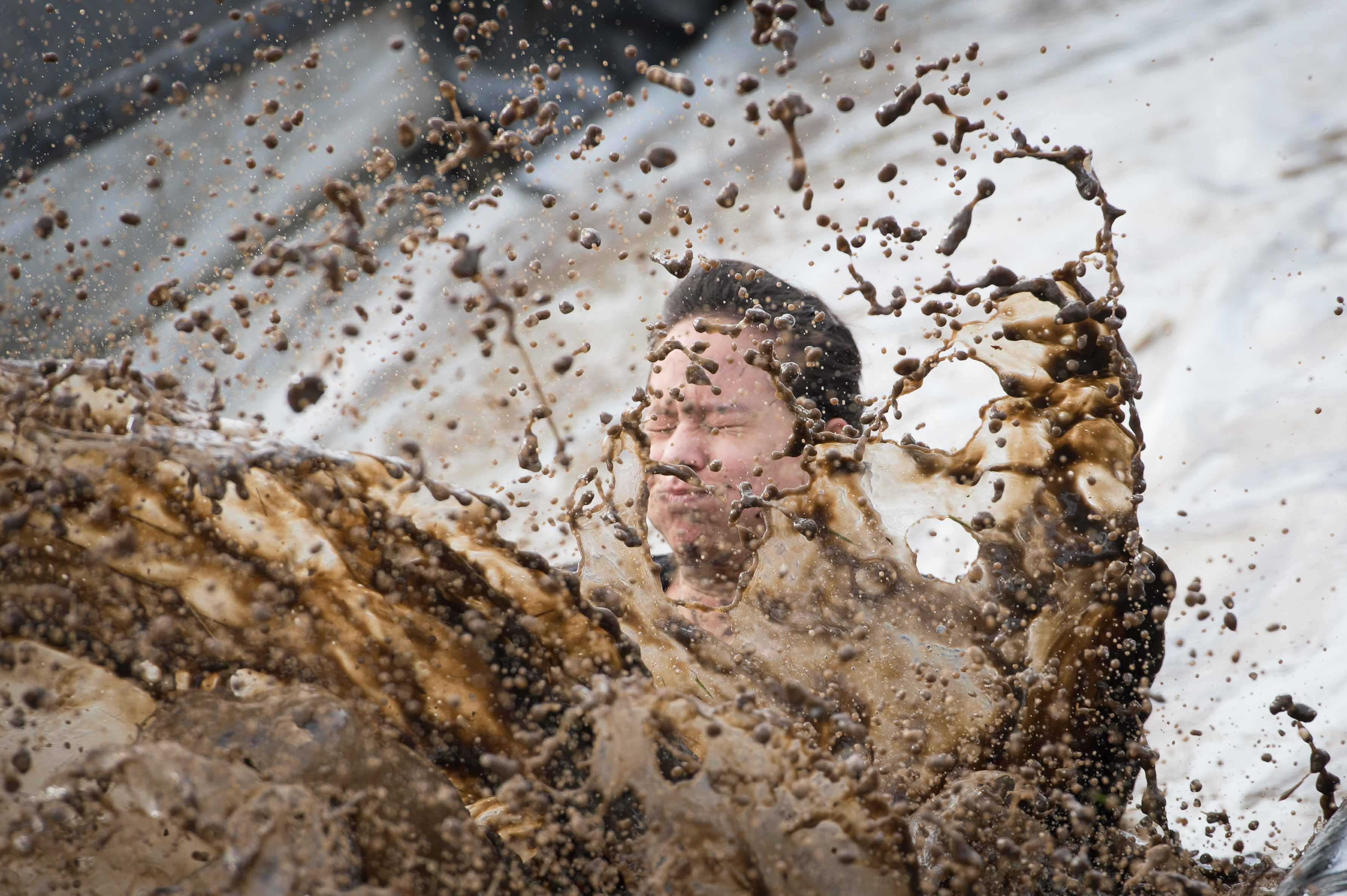 A man closes his eyes and holds his breath at the end of a large inflatable shoot makes a large muddy splash