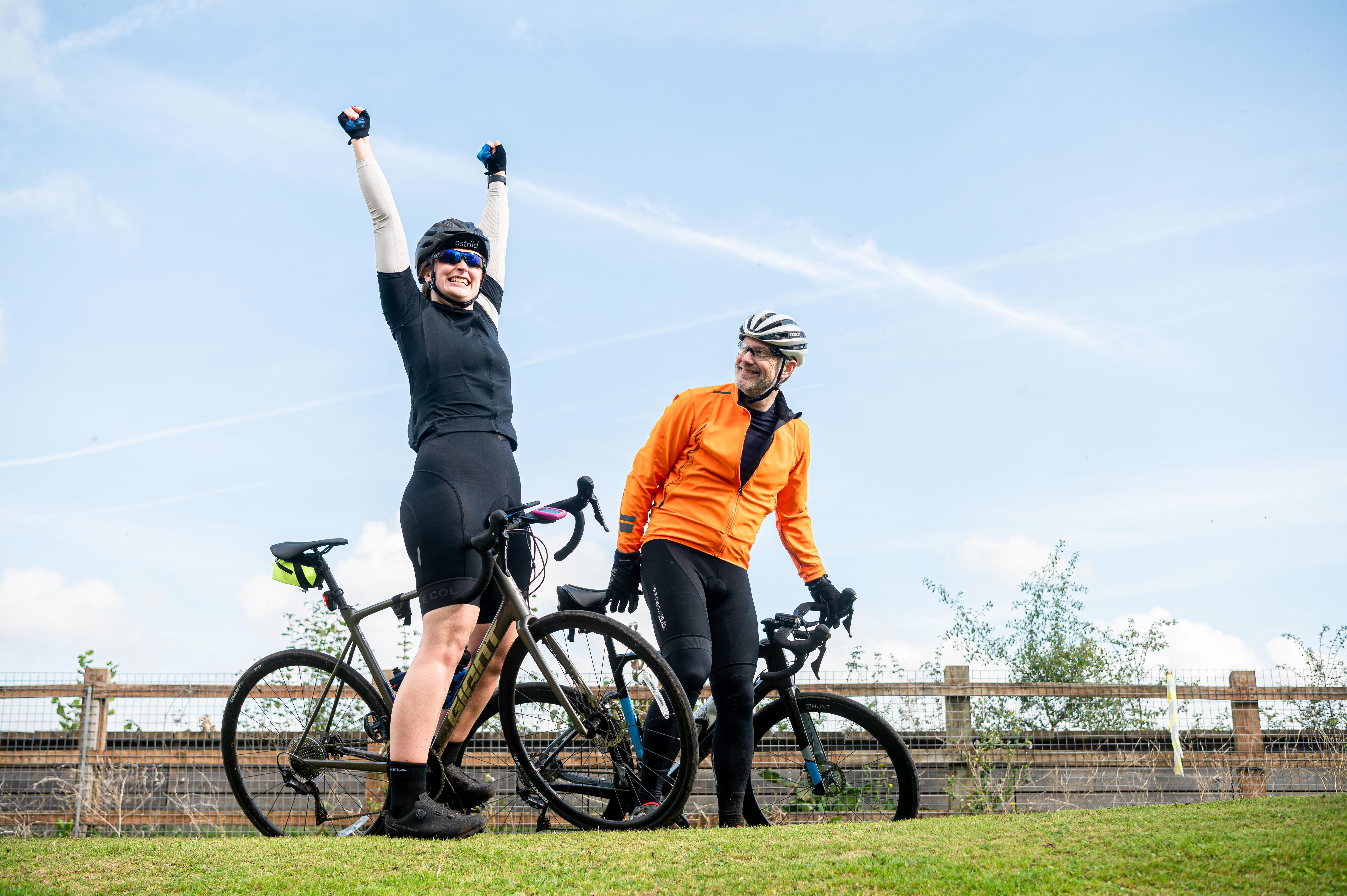 Two stationary cyclists are on an elevated grassy verge with a big blue sky. One is a woman smiling gleefully dressed in black with her arms vertical as if celebrating and the other cyclist is a man wearing an orange top and white helmet smiles at the other rider.