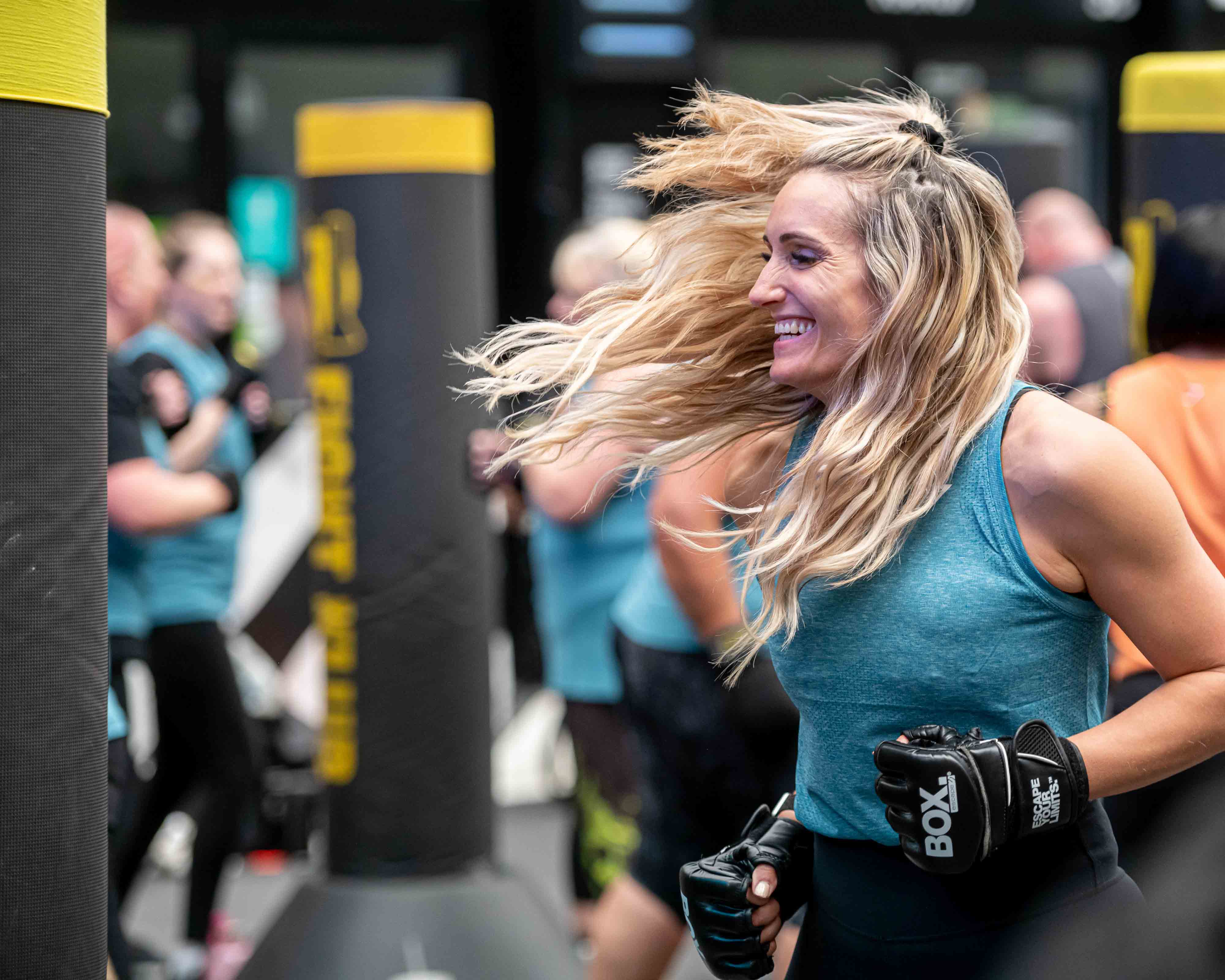 A woman dressed sports clothing and wearing boxing gloves smiles as she lunges back from hitt ing  a punch bag