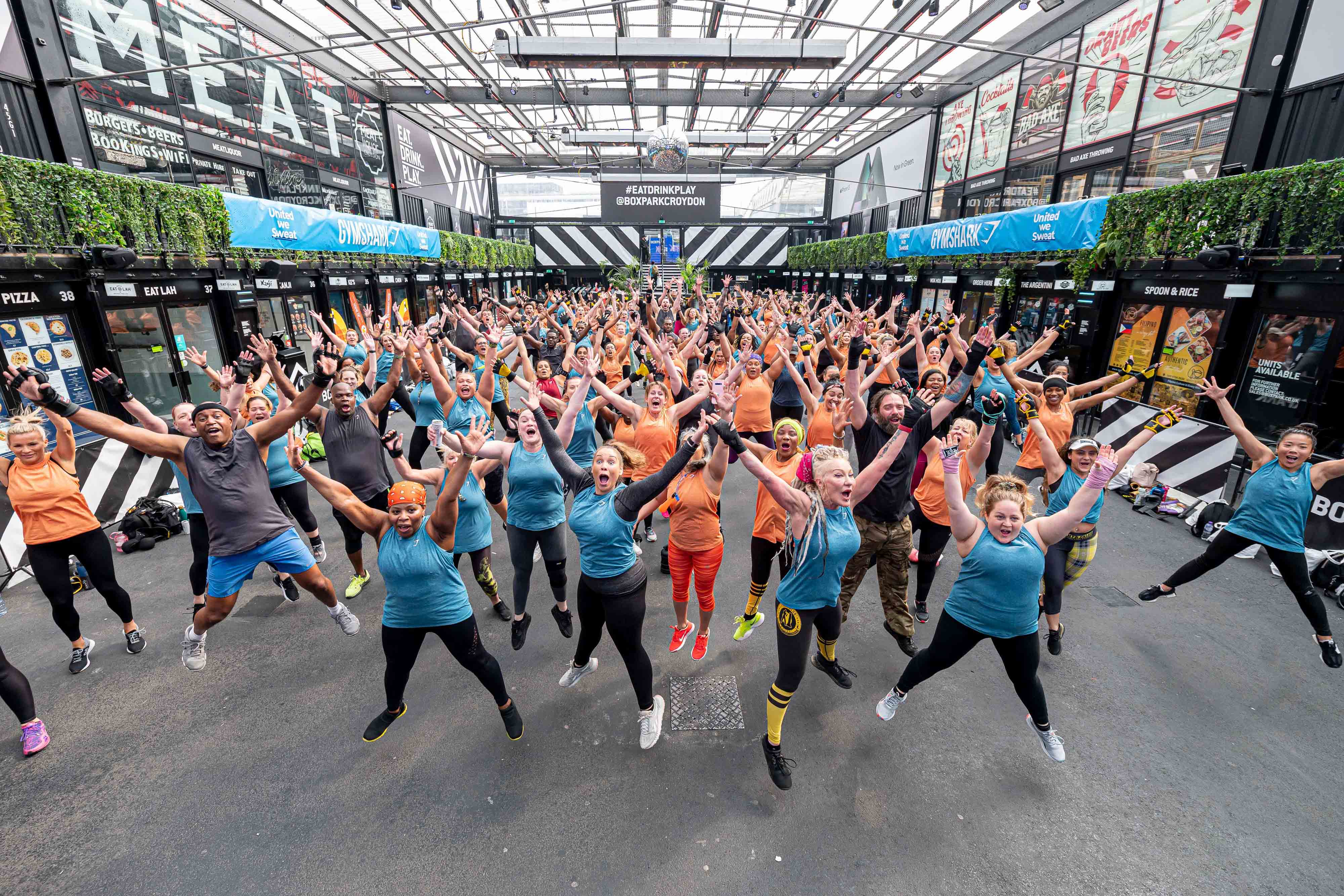 A large group of mostly women dressed in blue and orange vests all leap into star jump