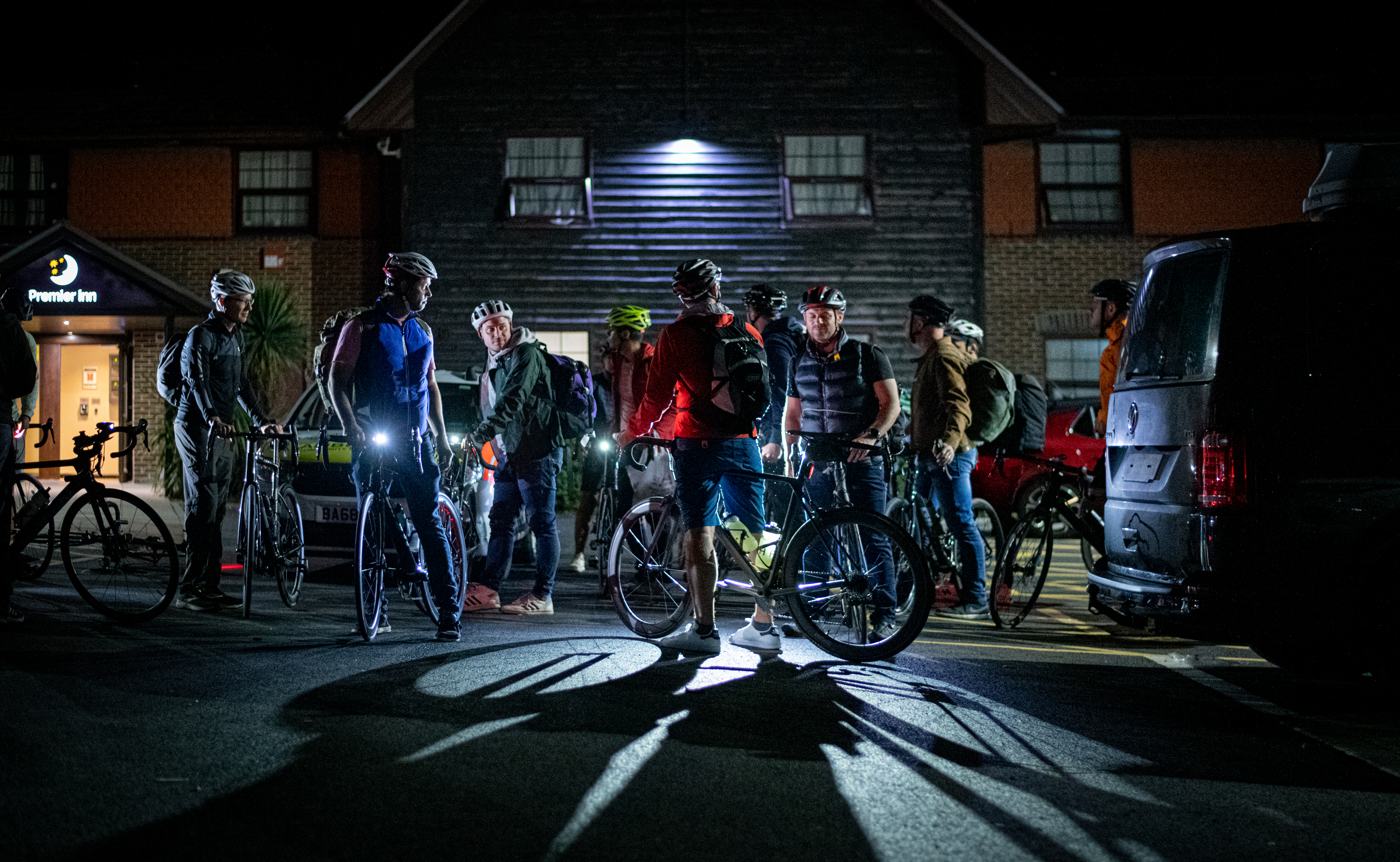 Cyclist grouped together outside a building in the dark ready to set off. A light behind them creates a dramatic shadow.