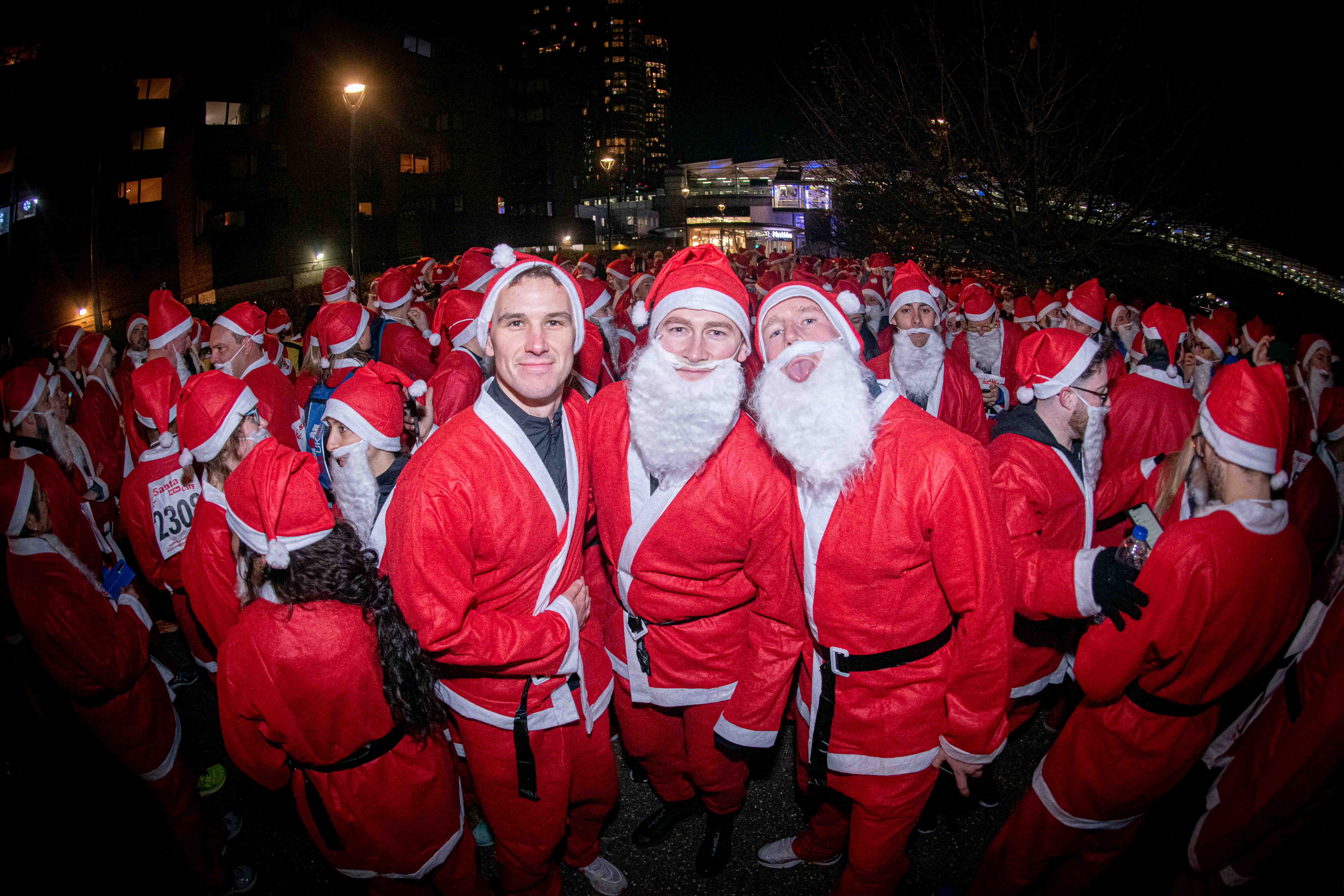An epic gathering of men and women dressed as Santa arrive at the start of a race in central London. Its night time and 3 men pose for a photo with the rest of the runners in the background.