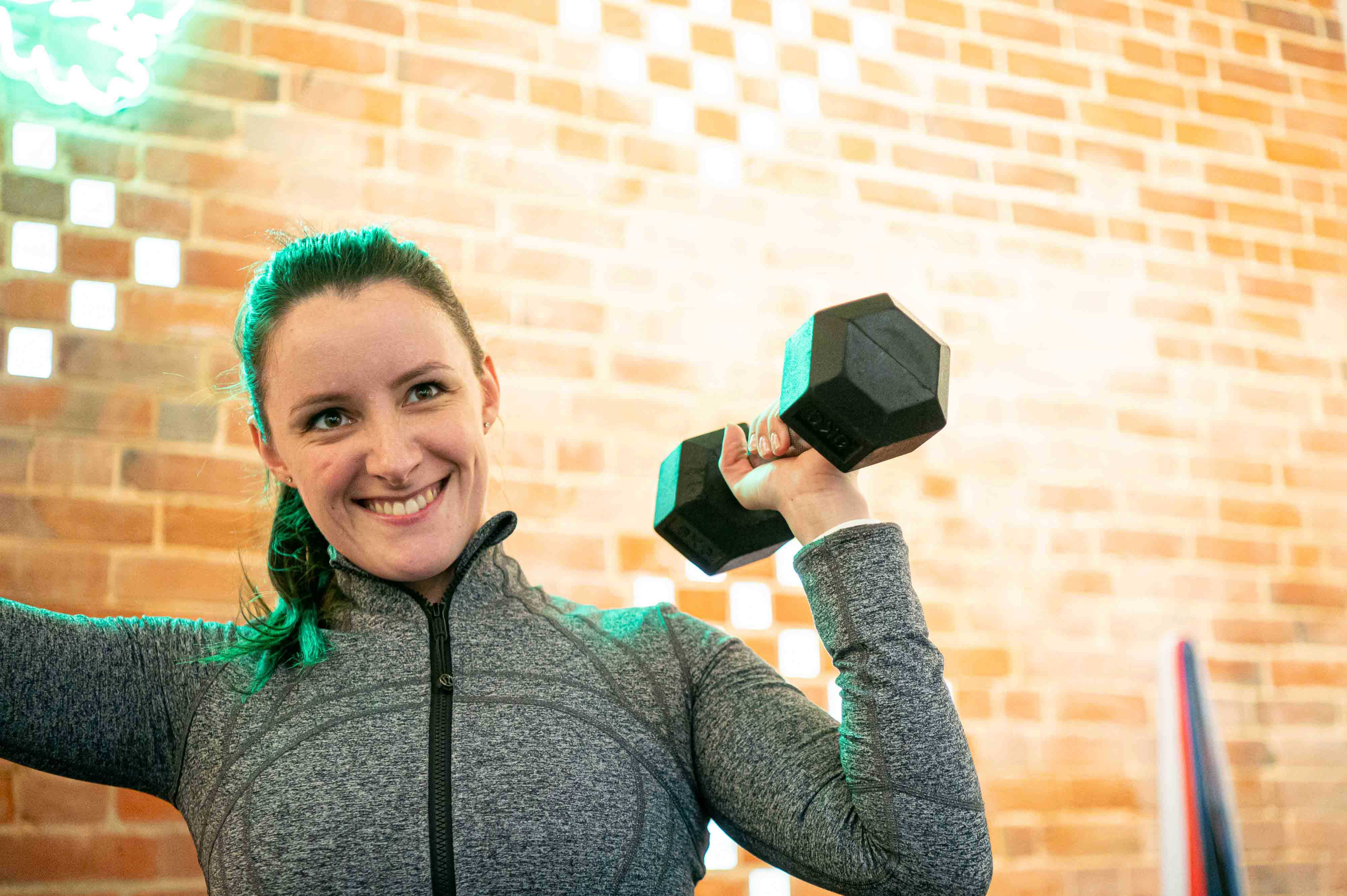 A woman in a grey gym top stands in front of a brick wall holding up a dumbbell. You can only see her left arm and top half as she smiles slightly off camera.