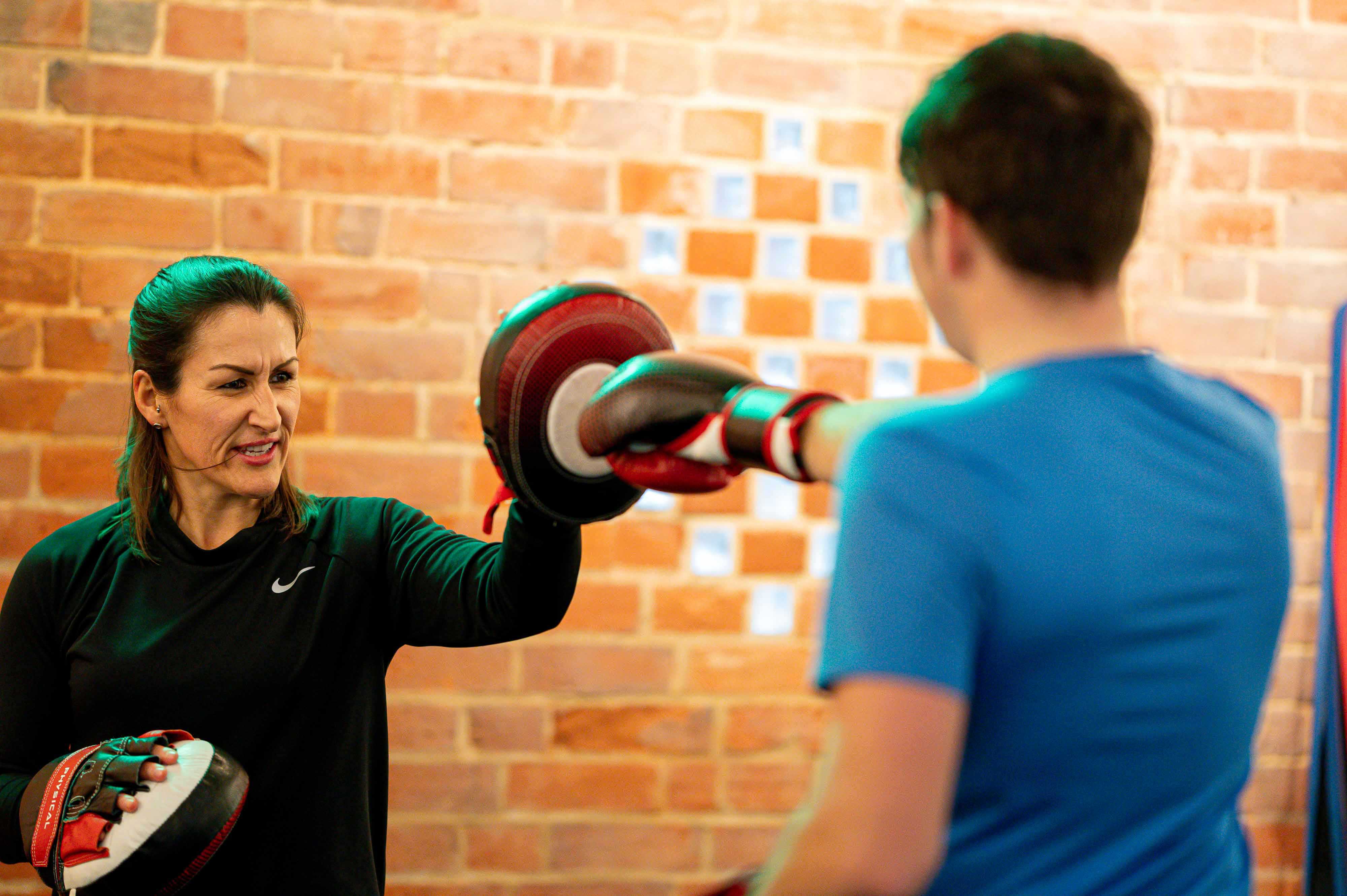 A female fitness instructor stands infant of a brick wall and is training a male client in boxing techniques as she holds boxing pads in front of him.
