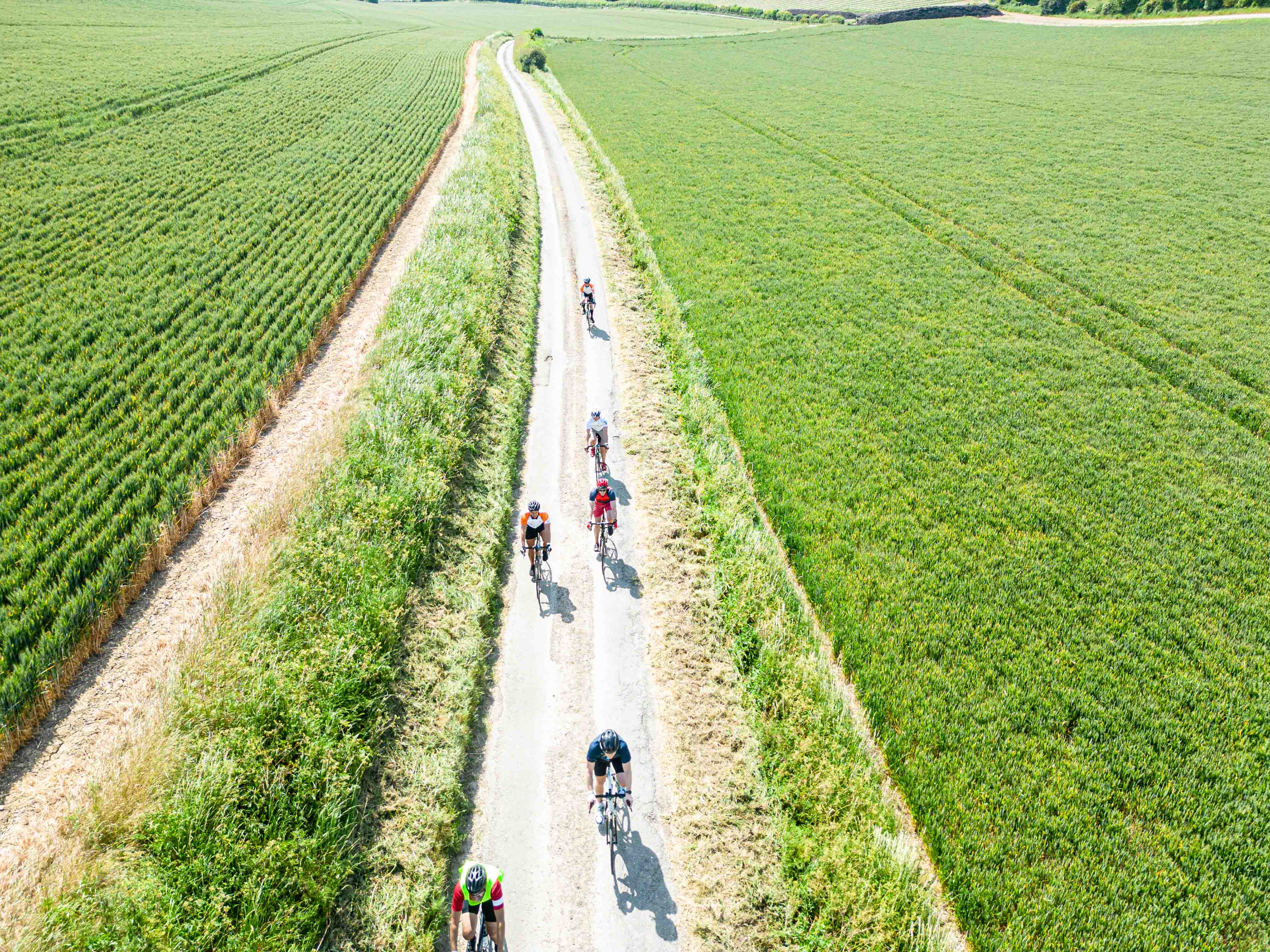 A group of cyclists travel an open country lane in the summer surrounded by green fields either side. The photo is taken from an elevated position looking down the road.