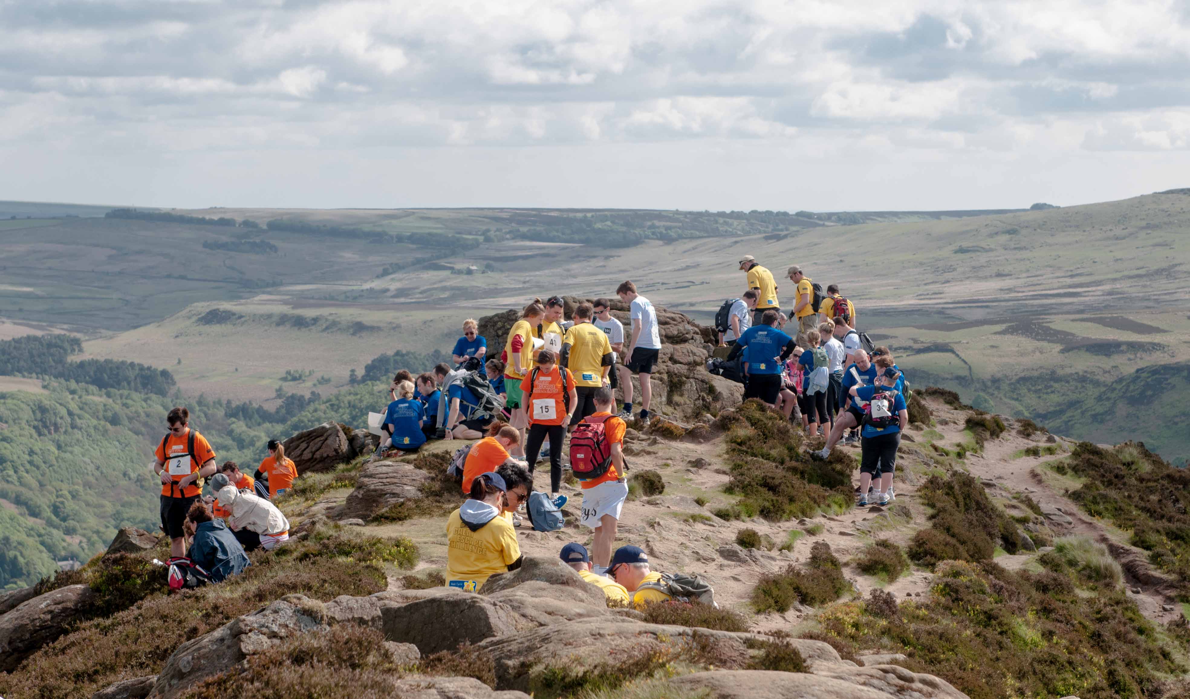 A large team of women and men look out over a spectacular view on a small mountain top covered in boulders.