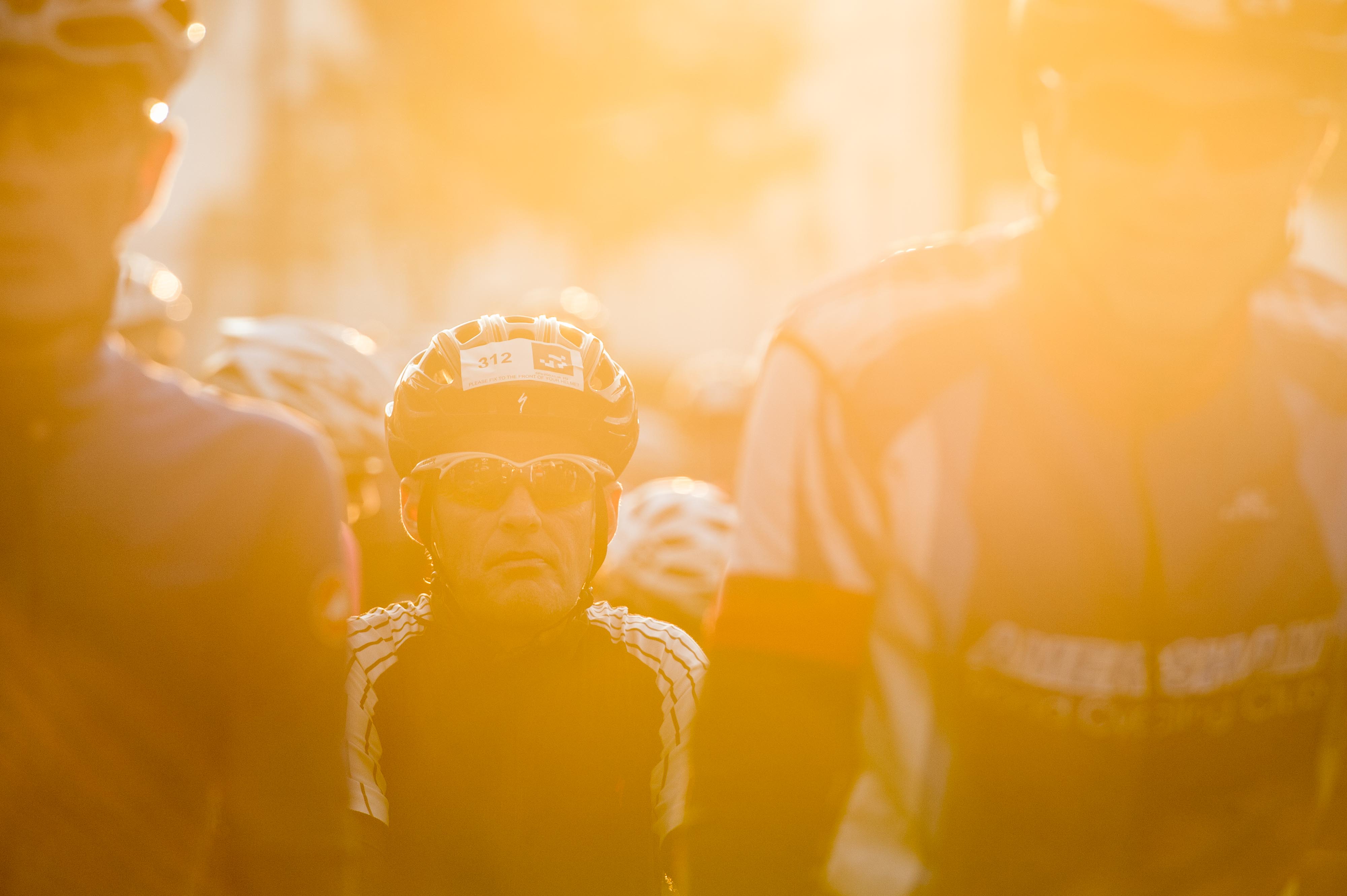 Cyclists preparing to set off in the morning sunlight. Close up of a man looking pensive, flanked by other blurred riders in helmets and jerseys.