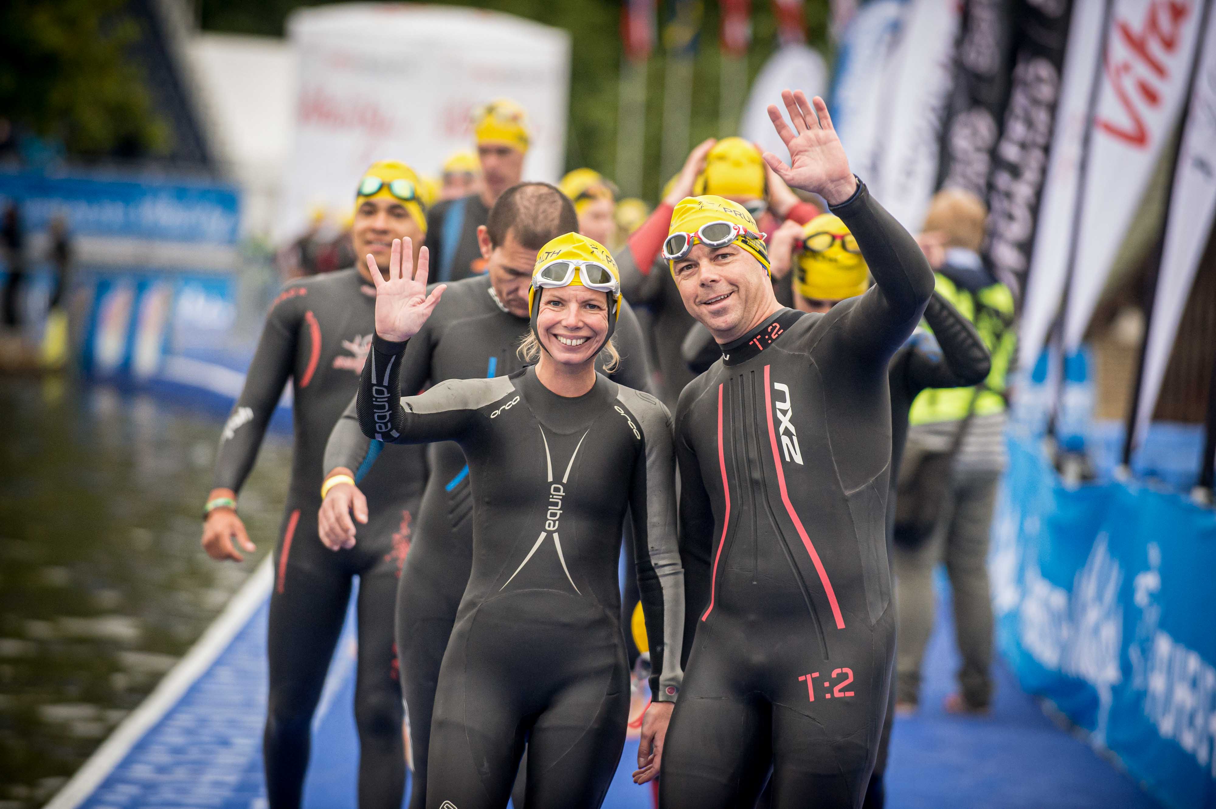 A wave of competitors all wearing black swim suits and yellow caps arrive on a pontoon for the start of the swim section of a triathlon. A woman and a man at the front smile and wave as they approach.