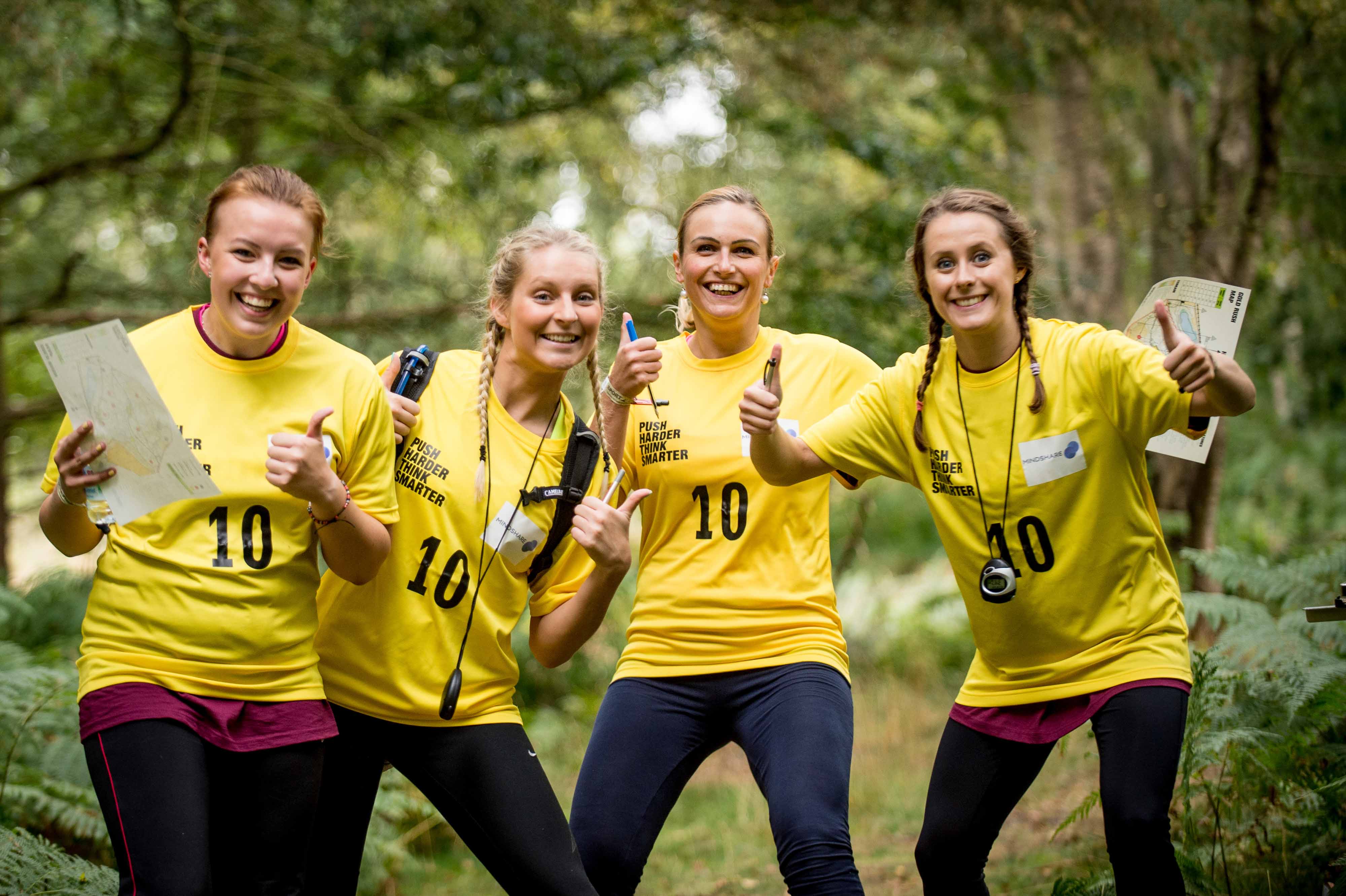 A team of four women outdoors in a wooded area smile and pose thumbs up for the camera